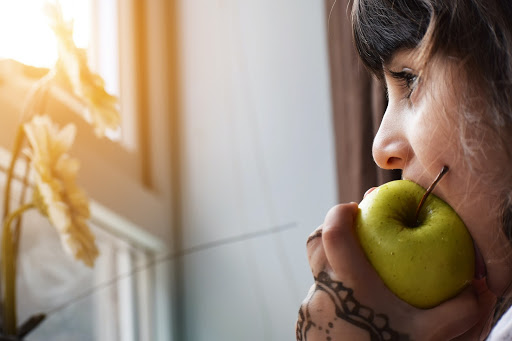 Person chewing on a green apple, looking outside a window.
